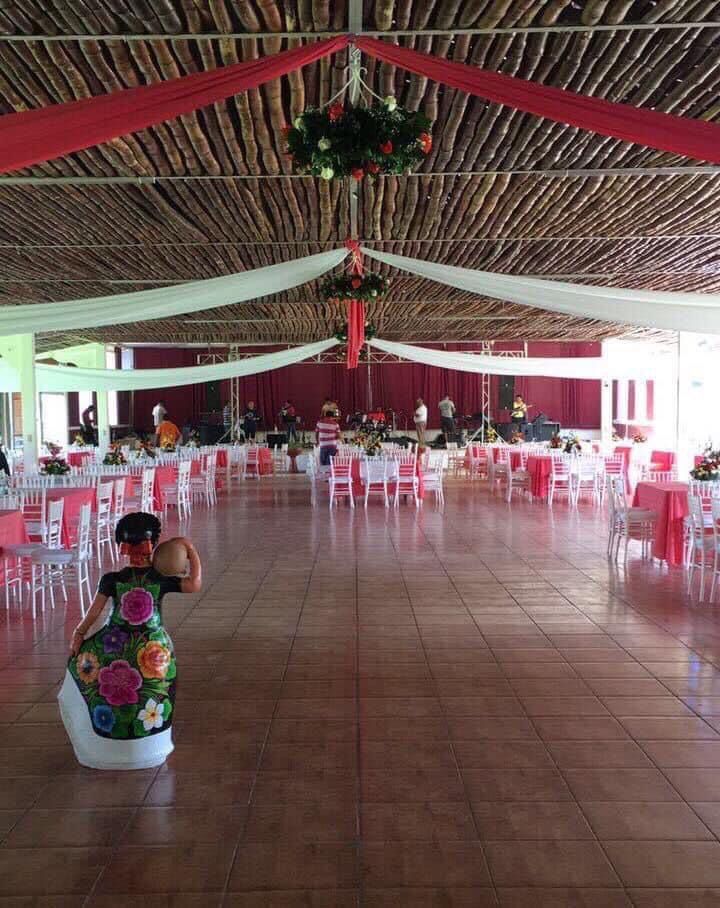 Salón de eventos con mesas preparadas para fiesta, decorado con tela roja y blanca, y un muñeco decorativo colorido en primer plano.