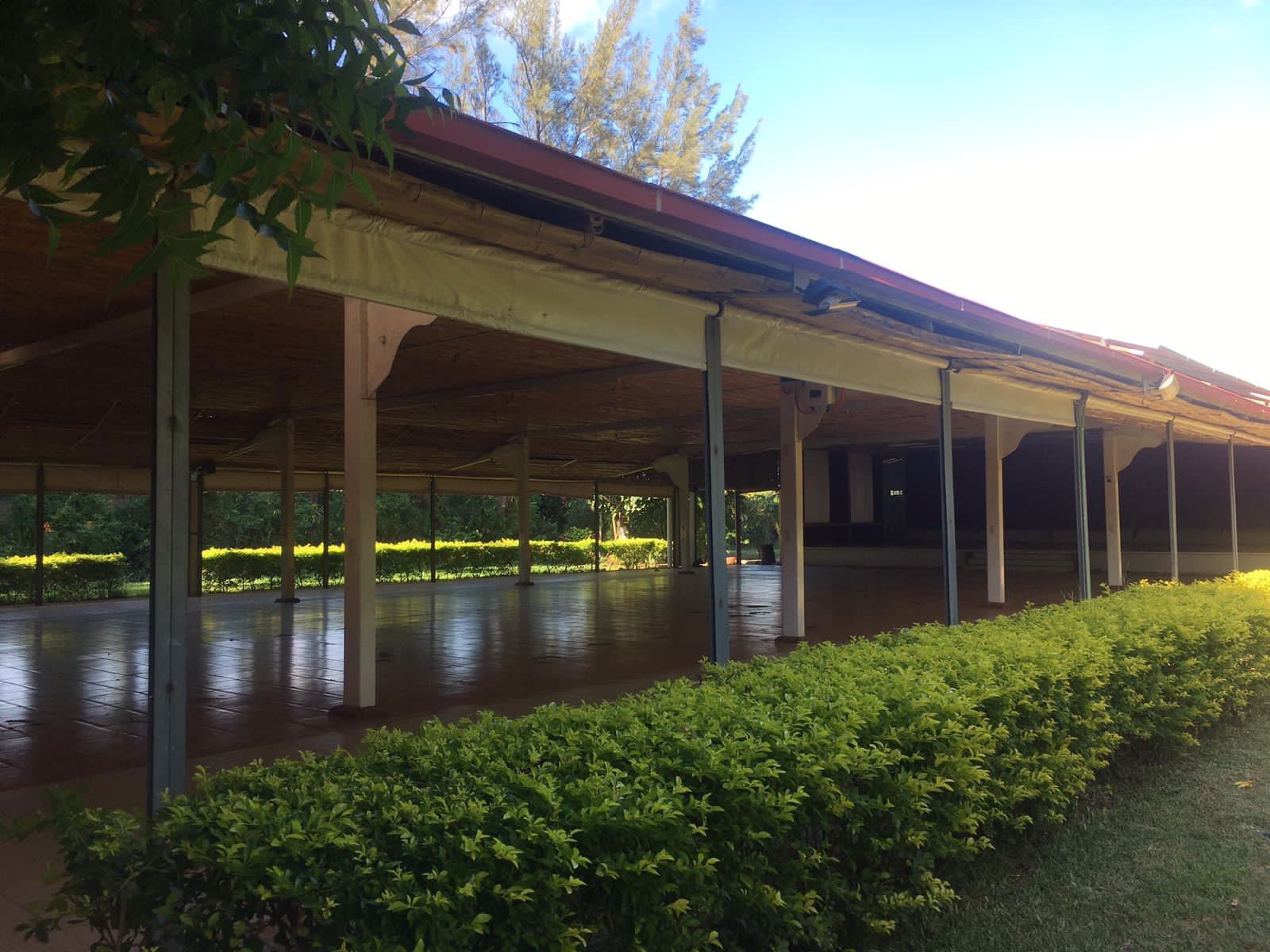 Pabellón al aire libre con techo rojo, sostenido por columnas blancas. Arbustos verdes bordean el primer plano.