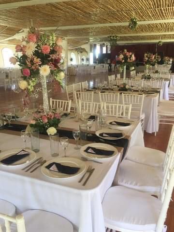 Salón de recepción con mesas decoradas, centros de mesa florales, sillas blancas y techo de bambú.