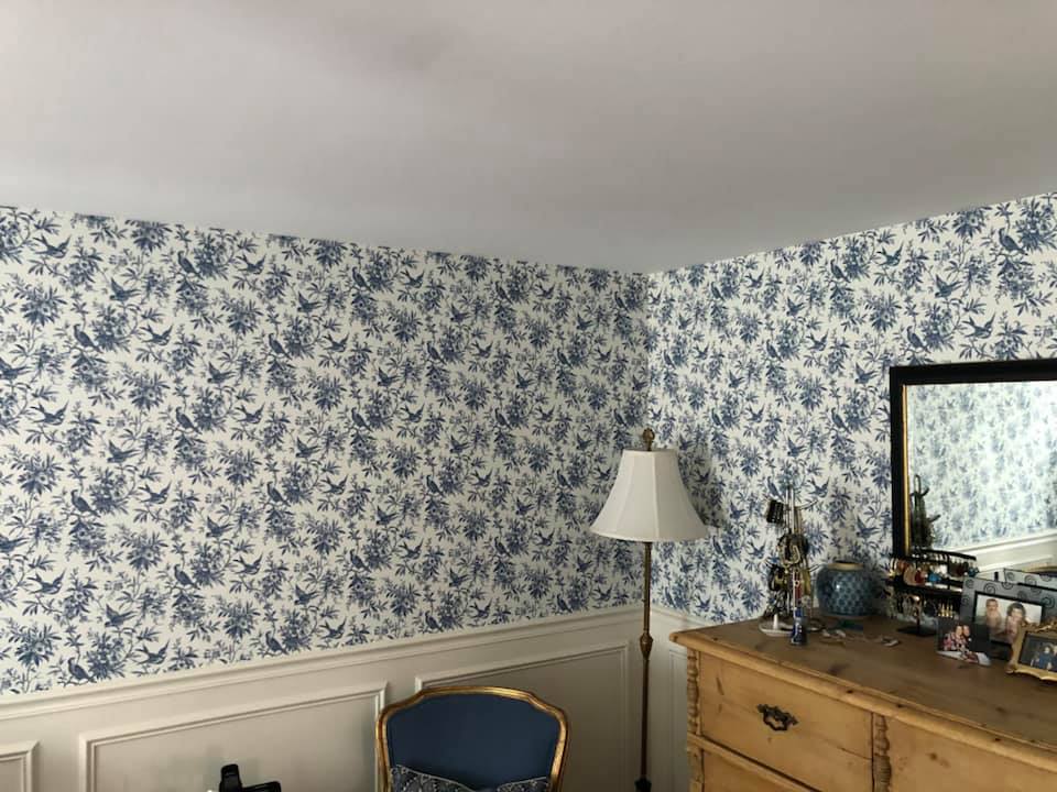 A bedroom with blue and white floral wallpaper , a lamp , dresser and mirror.