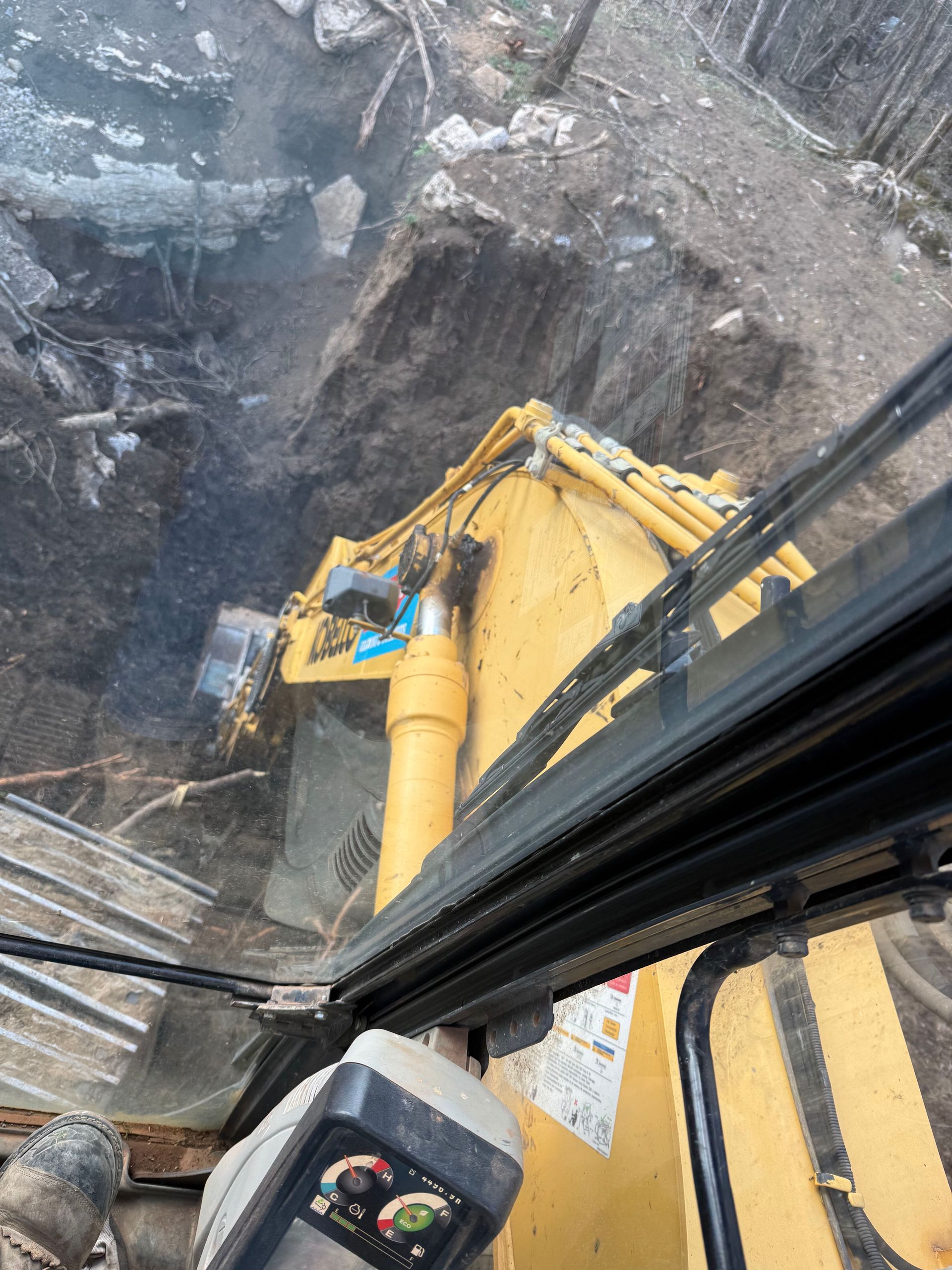 View from inside a yellow excavator cab looking down at a deep trench being dug in a wooded, rocky area.