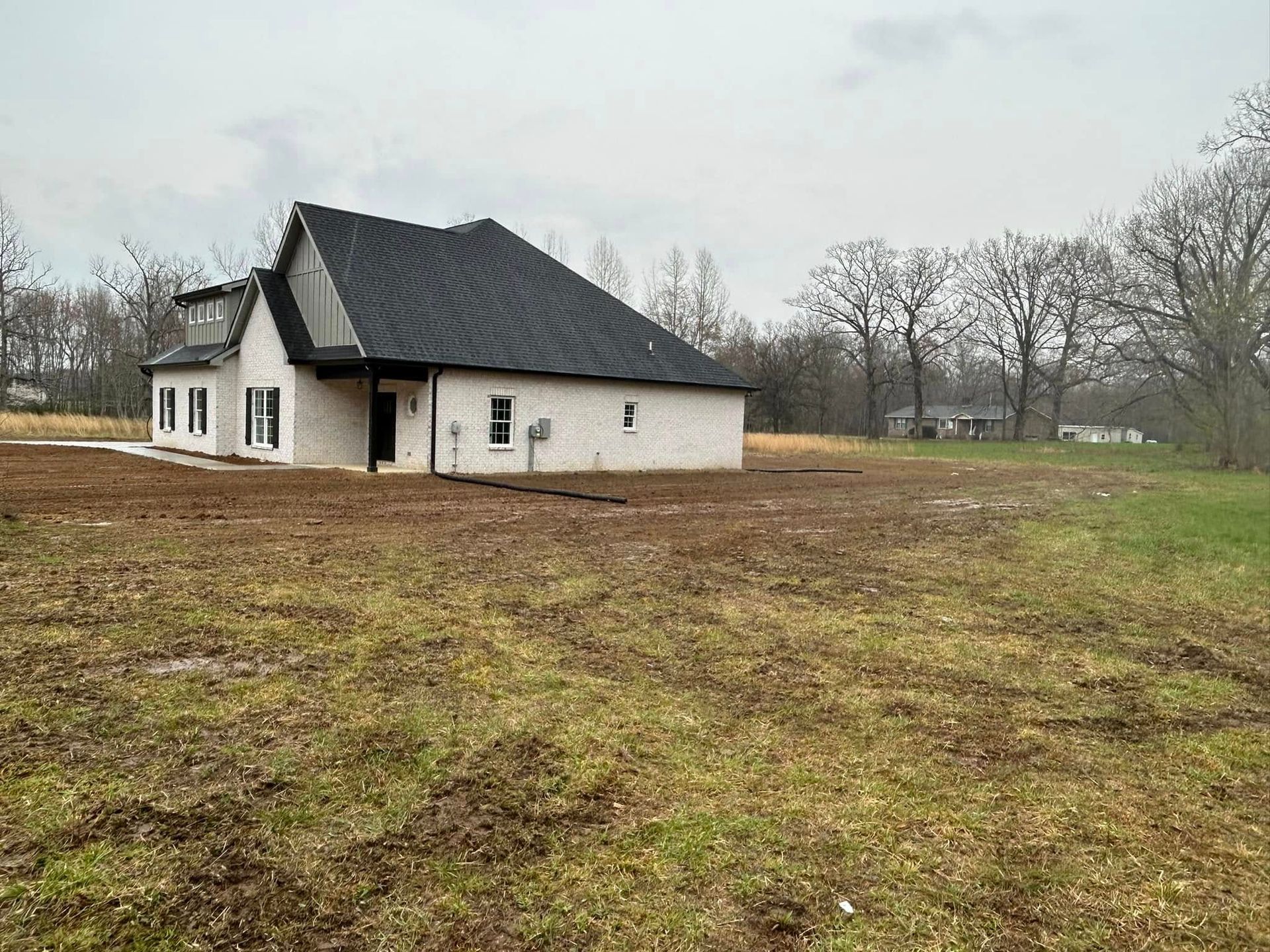 White brick house with black roof on a large, bare, brown field on an overcast day.