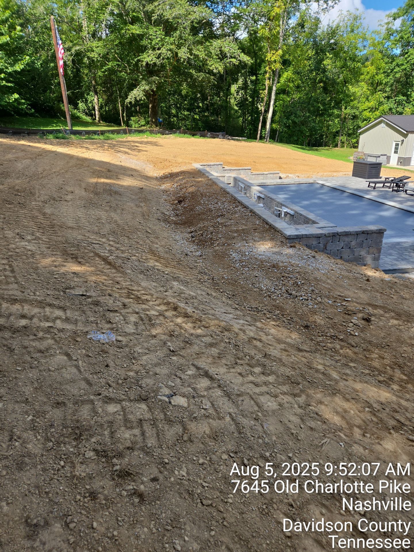 An outdoor view of a landscaped backyard with a stone pool deck under construction, surrounded by bare, graded dirt.