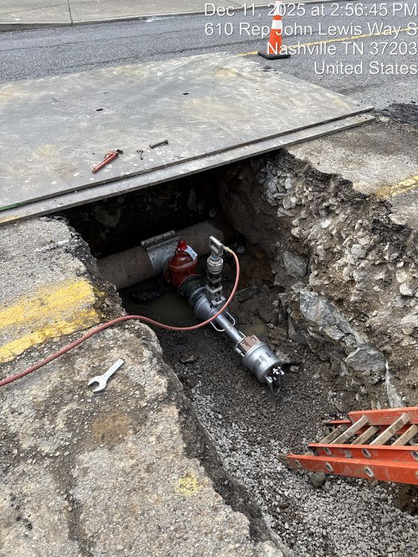 A construction trench in a street contains a pipe with a tapping machine attached, with a wrench nearby and a ladder.