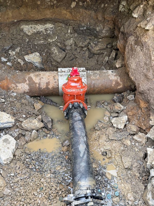 A red valve attached to a horizontal metal pipe, installed in an excavated dirt trench with surrounding rocks.