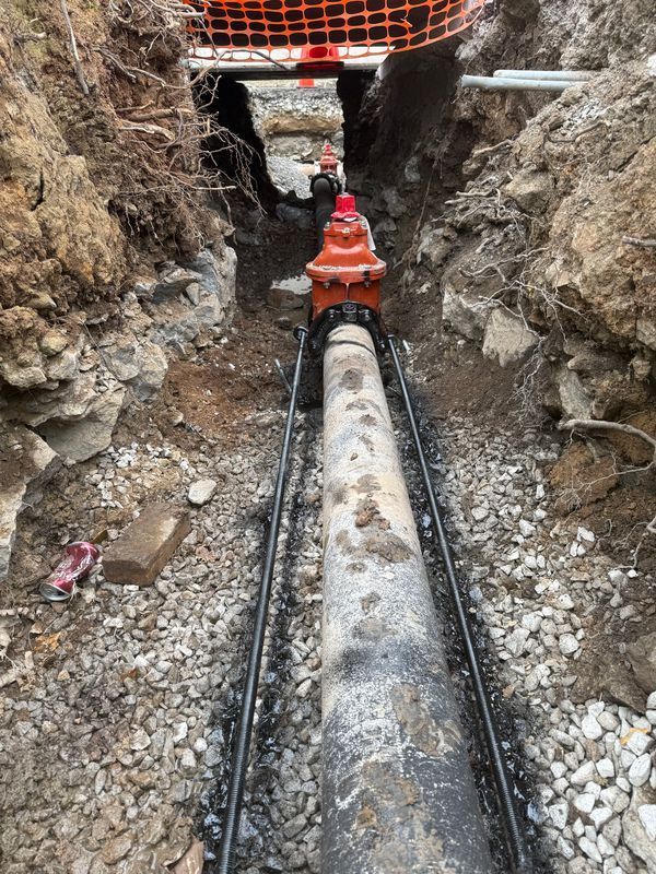A fire hydrant attached to a pipe in a narrow construction trench, surrounded by rocky soil and a protective orange fence.