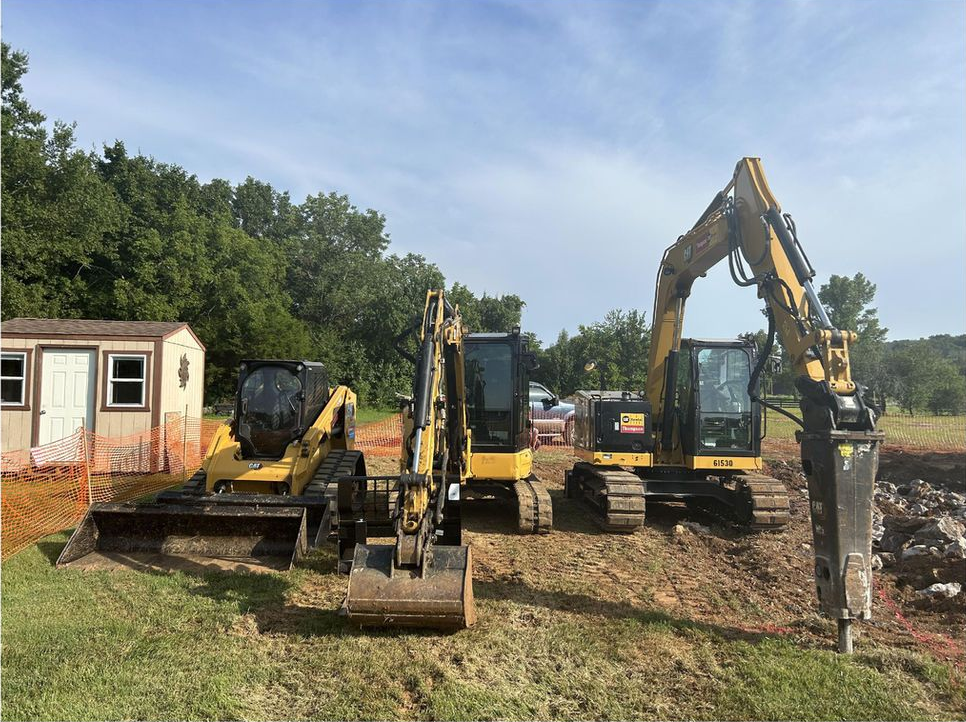 Three yellow excavators on a grassy patch near a small building and trees; some excavation work is underway.