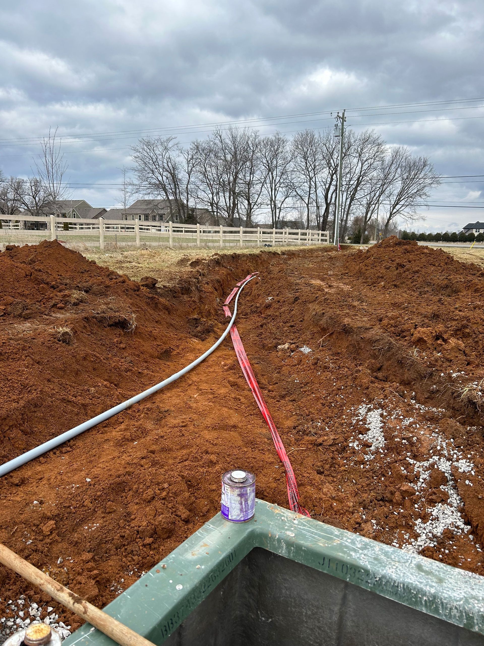 A trench in red soil with a conduit and red utility tape leading to an open green structure in the foreground.