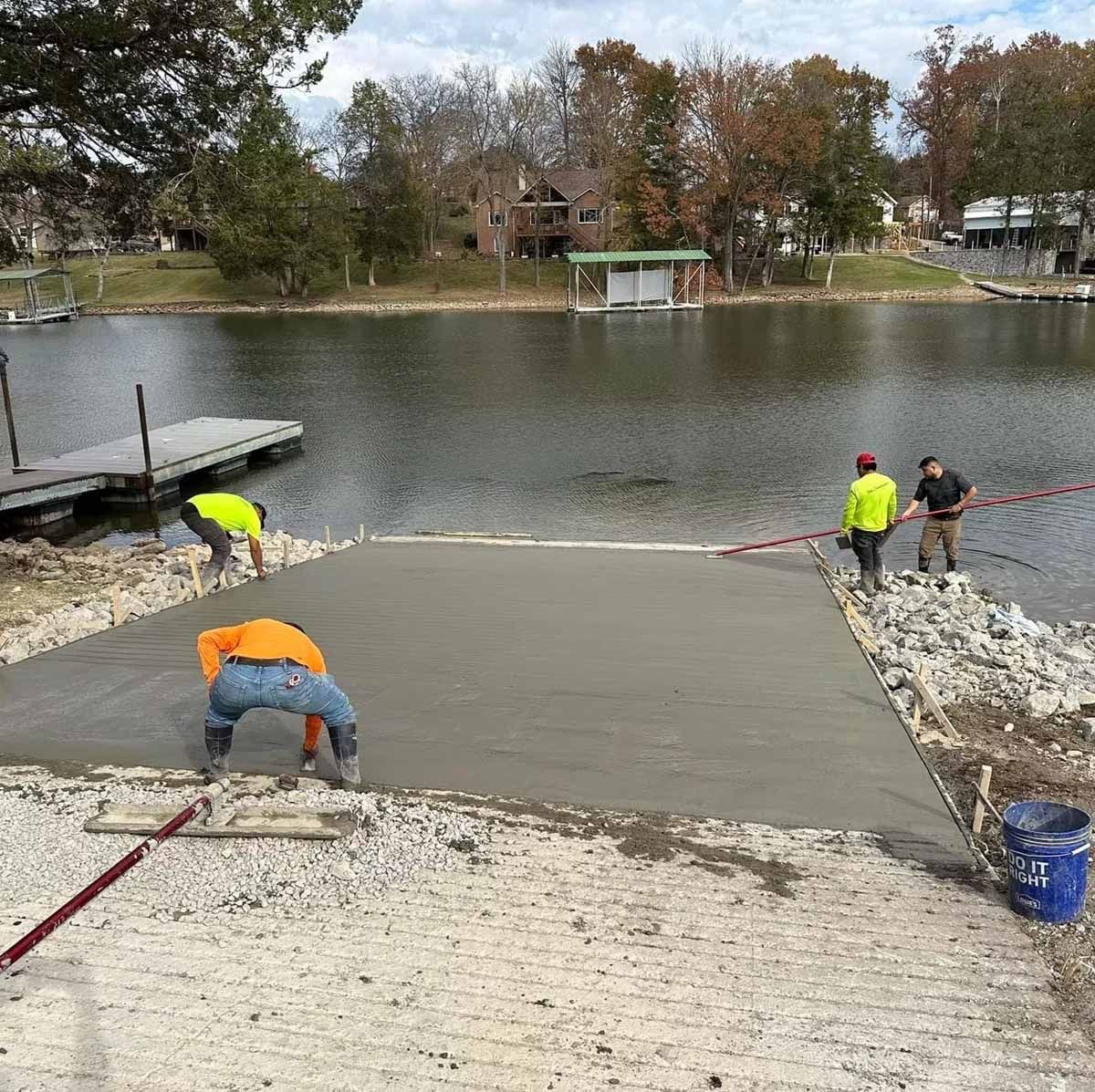 Construction workers pouring concrete for a boat ramp next to a lake with docks and houses.