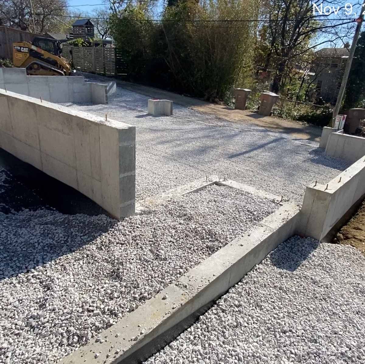 Construction site with concrete walls, gravel ground, and a small excavator in the background.