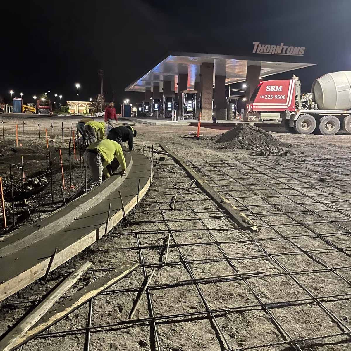 Construction workers pouring concrete curb at a Thornton's gas station at night. A cement truck is visible.