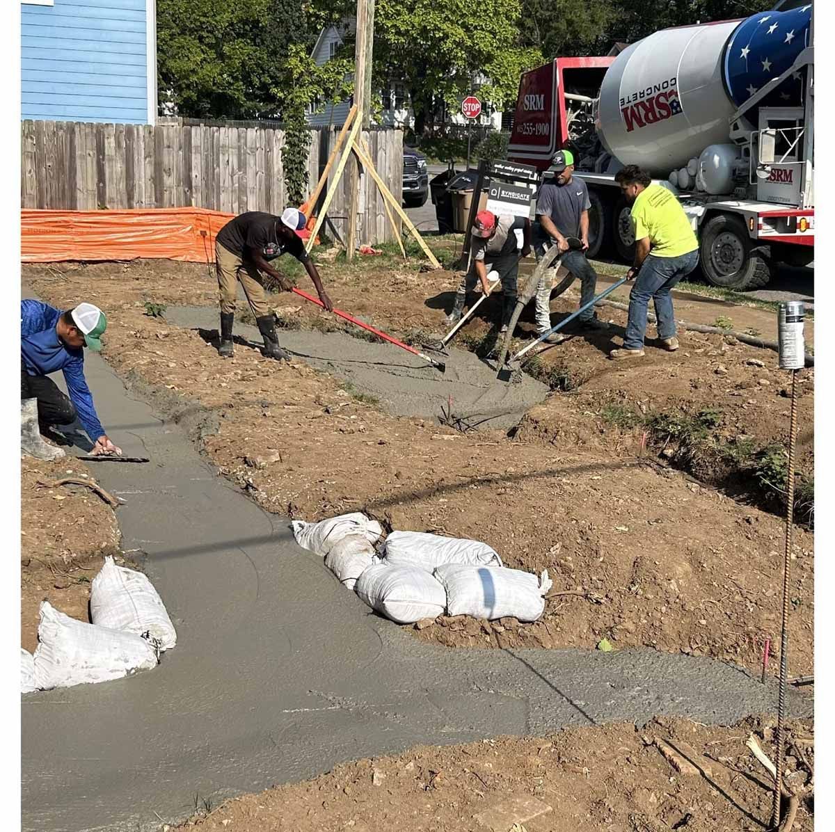 Construction workers pouring concrete for a sidewalk. A concrete mixer truck is in the background.