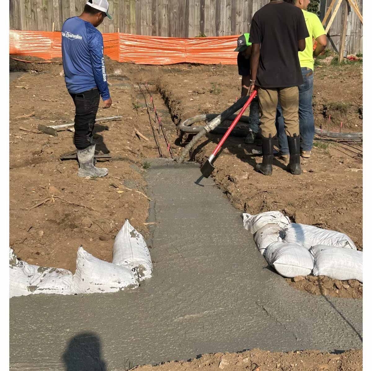Construction workers pour concrete for a foundation. Men are using tools on a dirt lot, with sandbags and rebar visible.