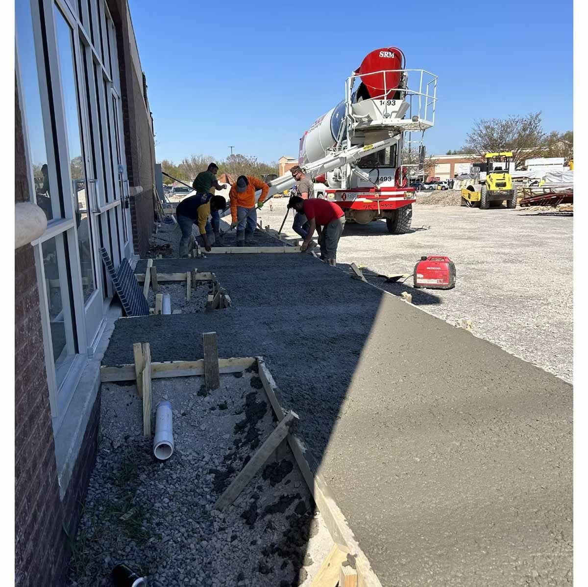Construction workers pouring concrete near a building, with a cement truck and forms visible.