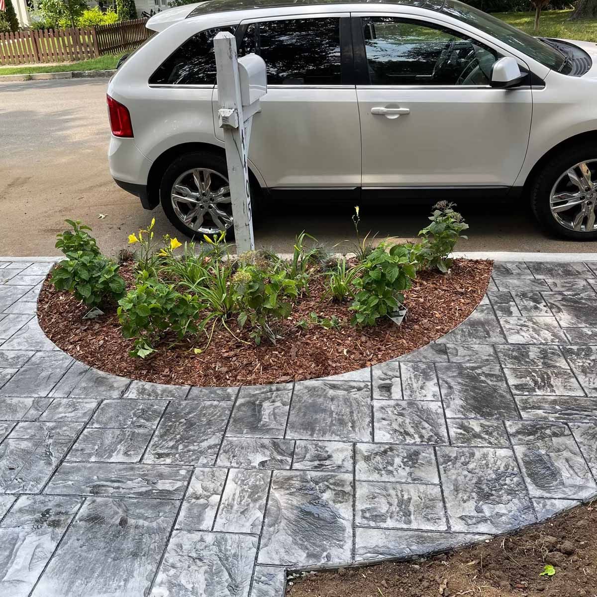 White SUV parked next to a grey patterned concrete walkway and small garden bed with plants.