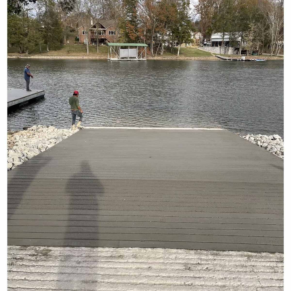 Boat ramp leading to a lake, with two people fishing off a dock. Brown buildings in background.