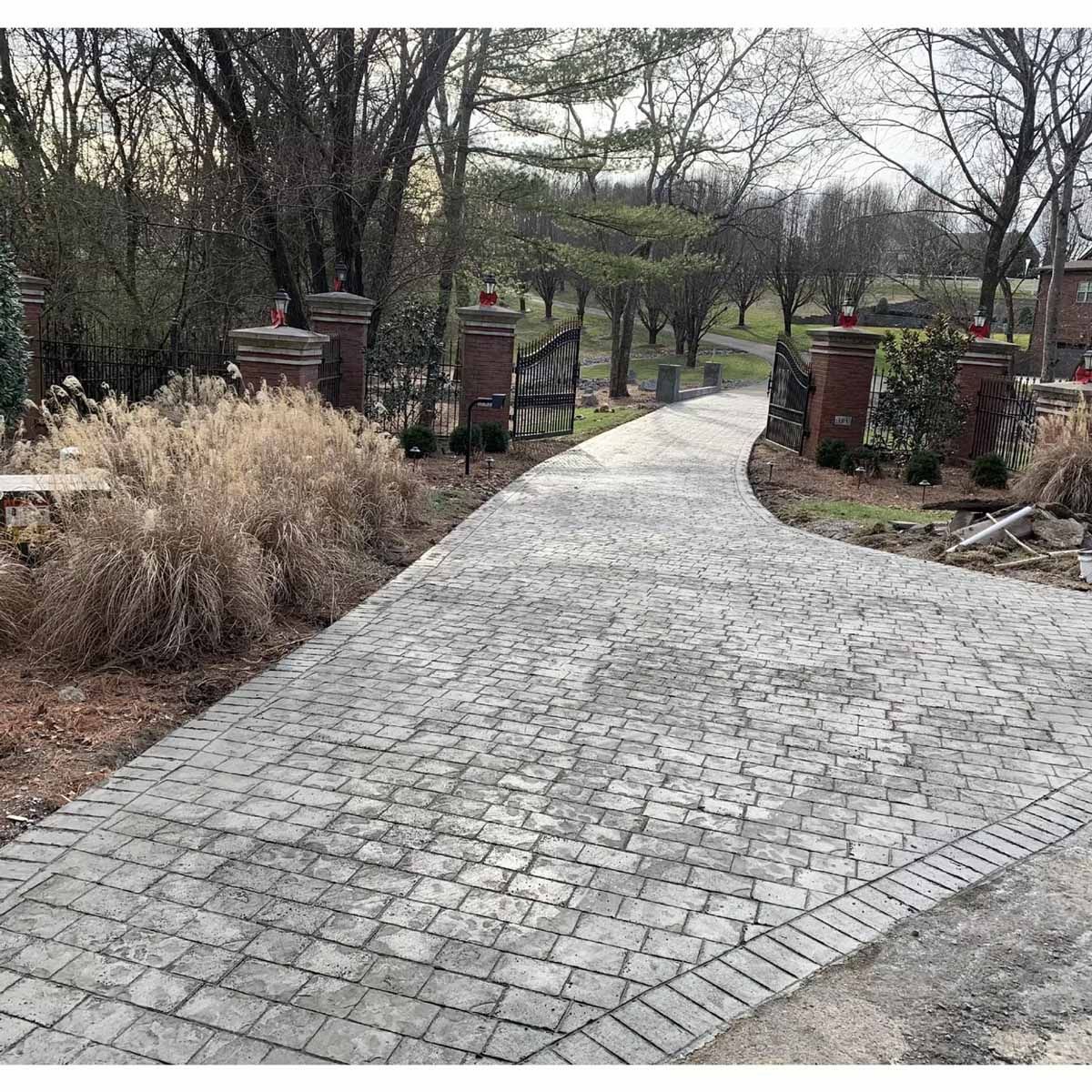 A paved driveway curves toward a wrought iron gate, flanked by brick pillars. Overcast, outdoor scene.