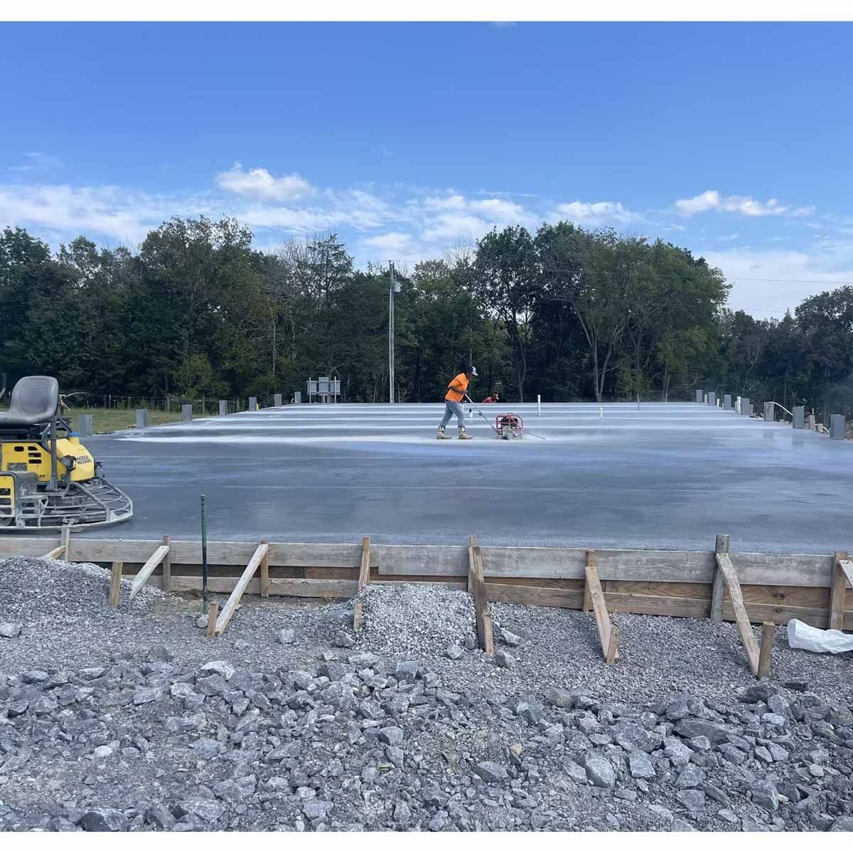 Construction worker finishing concrete slab on a sunny day. Gravel and wooden forms in foreground.