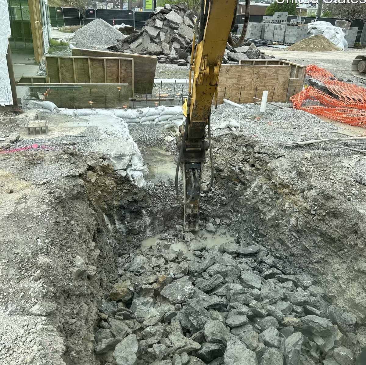 Excavator breaking up rocks in a construction site, dirt and debris visible.