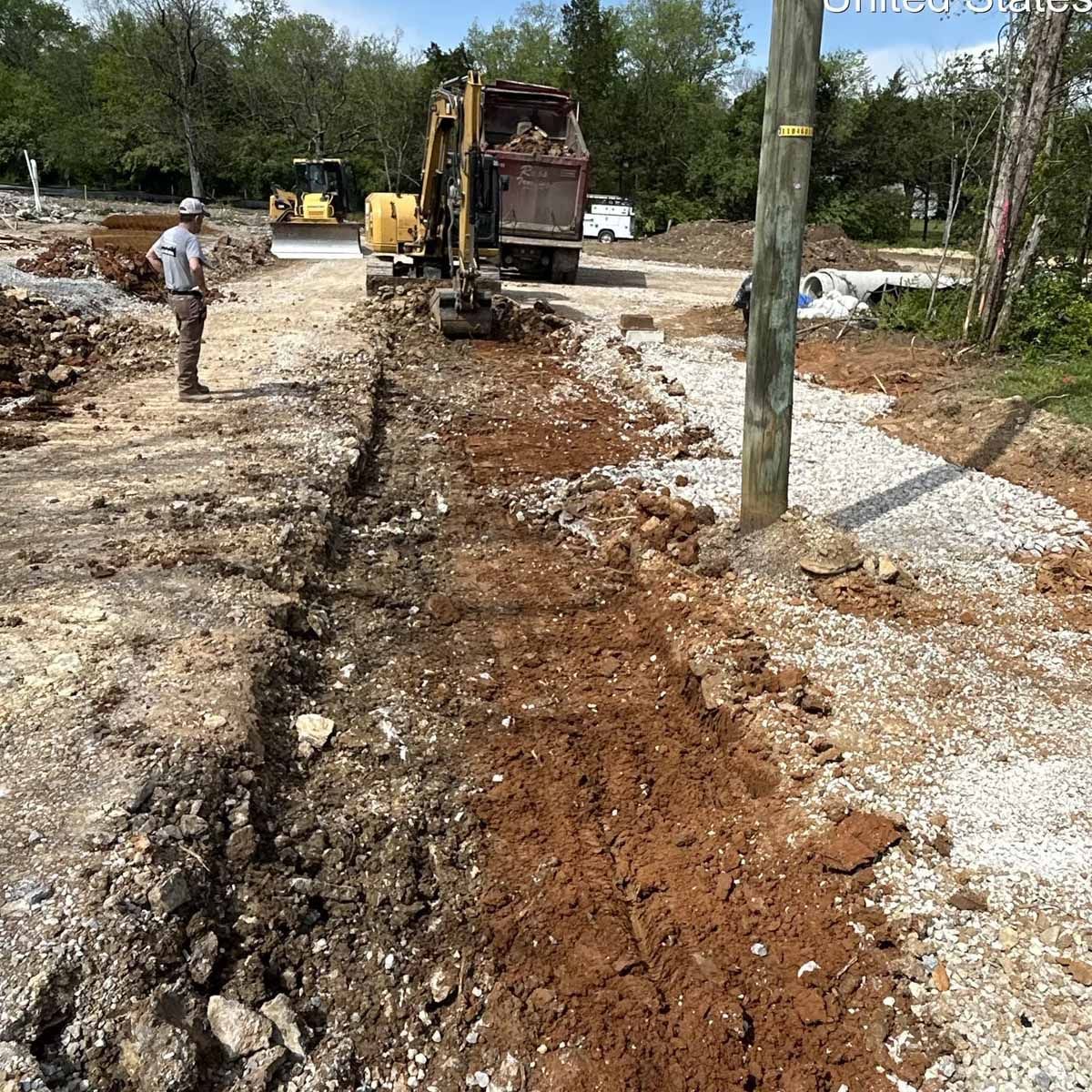 Construction site: Excavator digging trench, worker observing, dirt and rocks, sunny day.