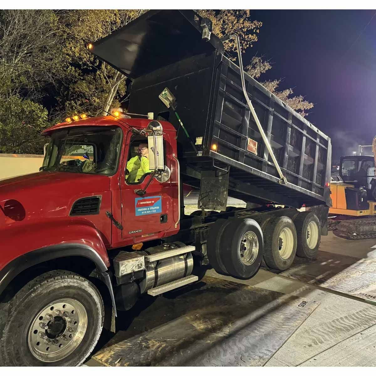 Red dump truck with raised bed, driver visible, during road paving at night.