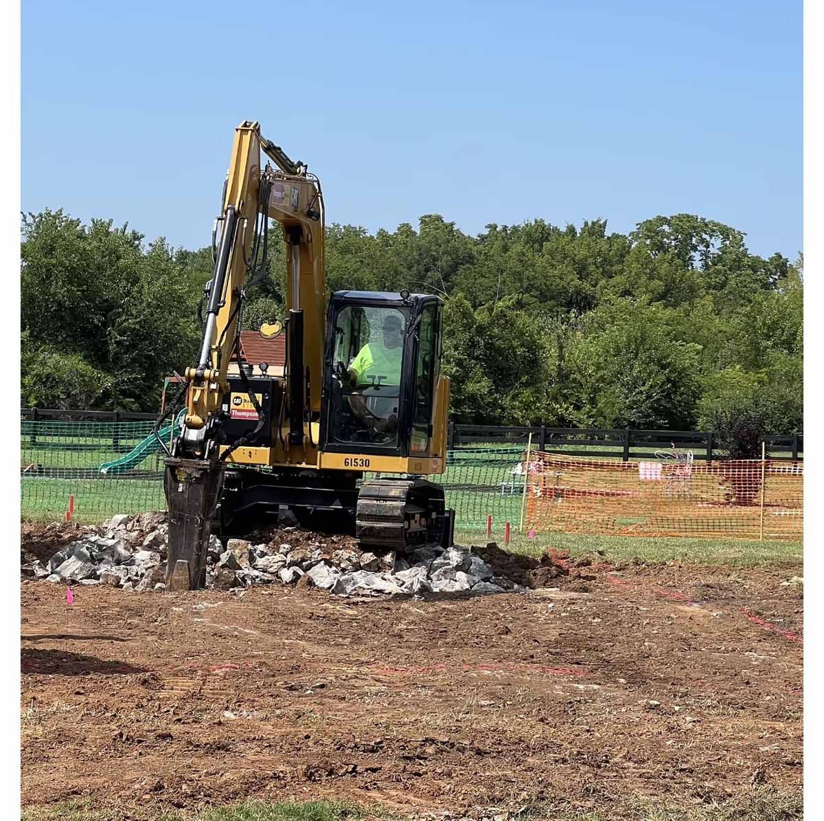 Yellow excavator demolishing debris in a construction site with a person inside; trees in background.