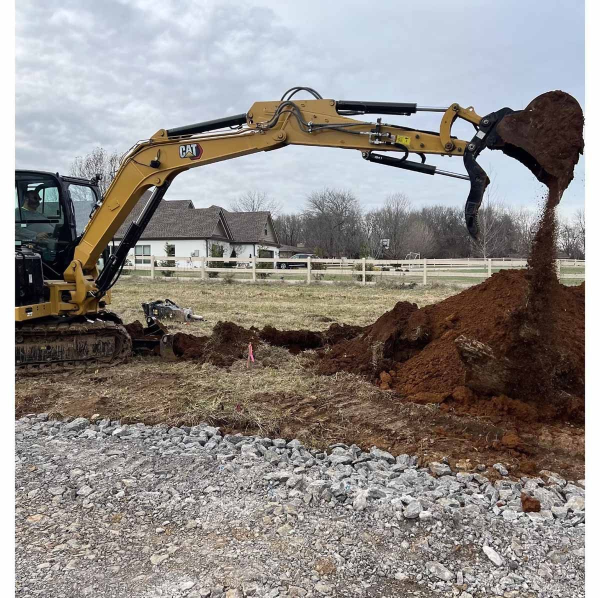 Yellow excavator digging soil in a field near a house and fence.