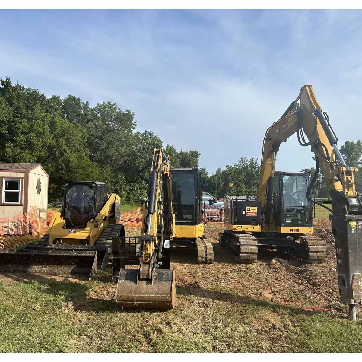 Three yellow excavators on a construction site with a small building, trees, and a blue sky.