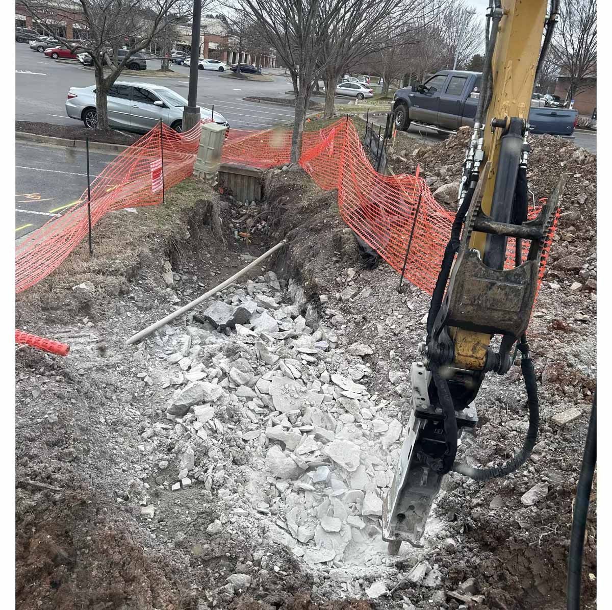 Excavator breaking concrete in a construction site. Orange safety fencing surrounds the pit.