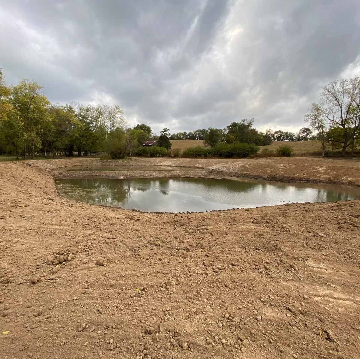 A newly dug pond with earthen banks reflecting a cloudy sky, surrounded by trees.