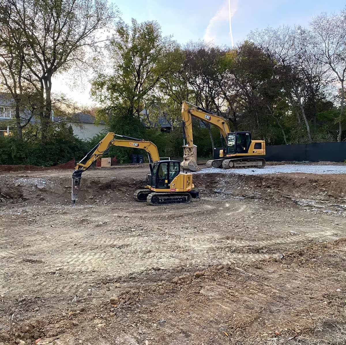 Two yellow excavators on a construction site, digging into the dirt. Trees and a building are in the background.
