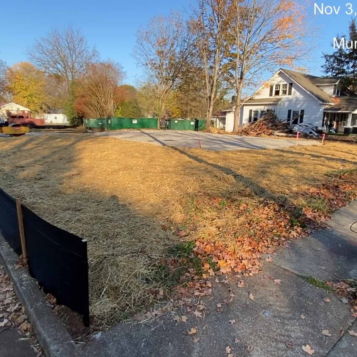 A vacant lot with dry grass, fenced off for construction. A house is in the background.