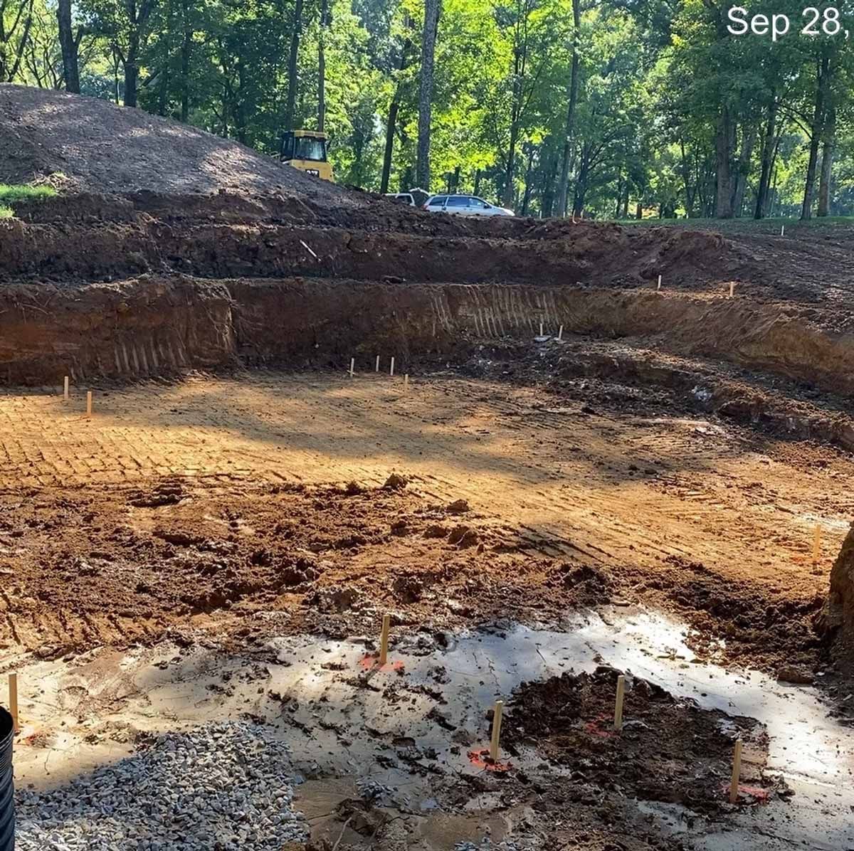Construction site with dirt, a muddy area, and a pile of earth, surrounded by trees.