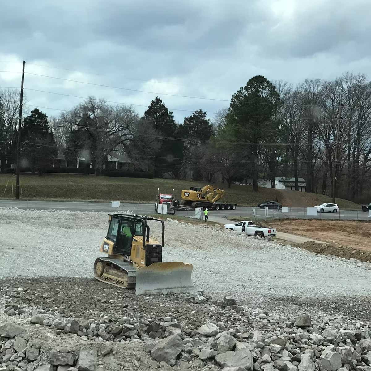 Bulldozer on construction site, preparing ground with gravel. Trees and road in background.