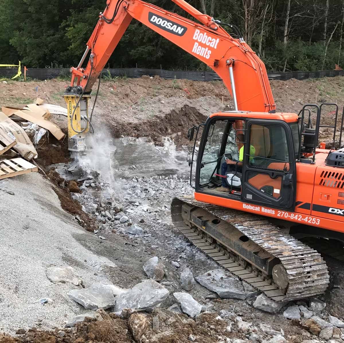 An orange excavator with a jackhammer demolishes concrete in a construction site.