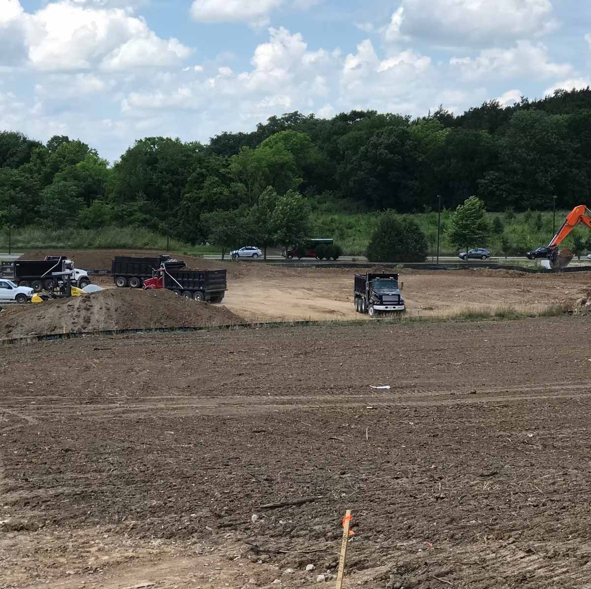 Construction site with trucks, excavators, and dirt under a partly cloudy sky, trees in the background.