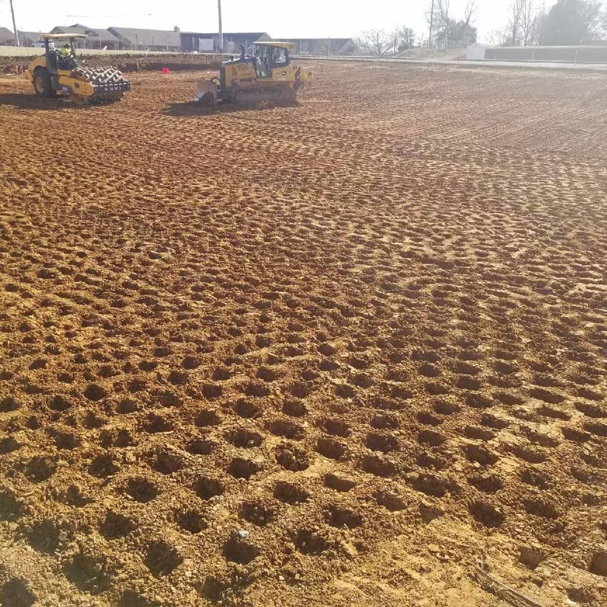 Construction site with soil marked with evenly spaced holes, heavy machinery in the background.