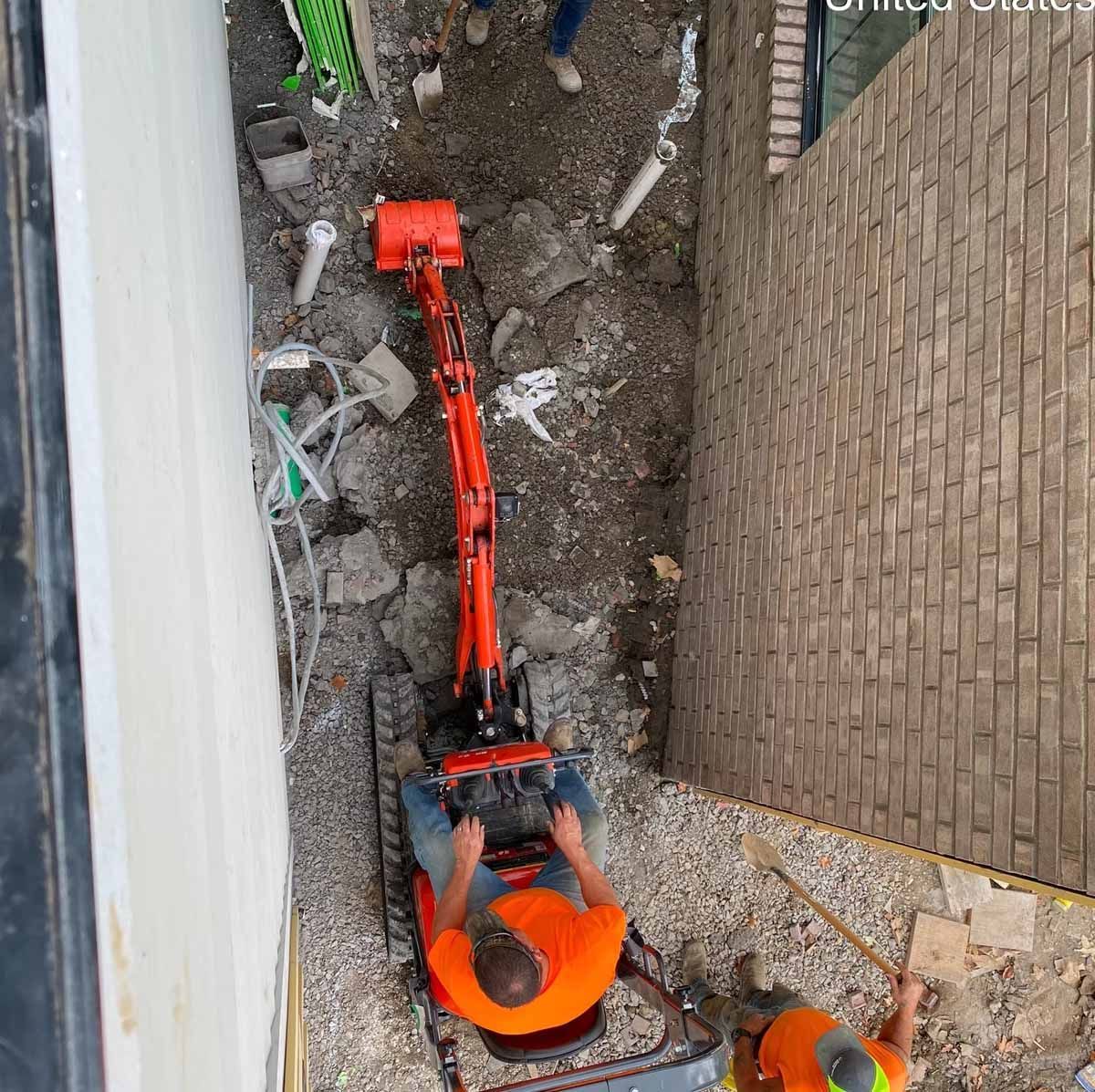 Construction workers using a small excavator to dig alongside a brick wall.