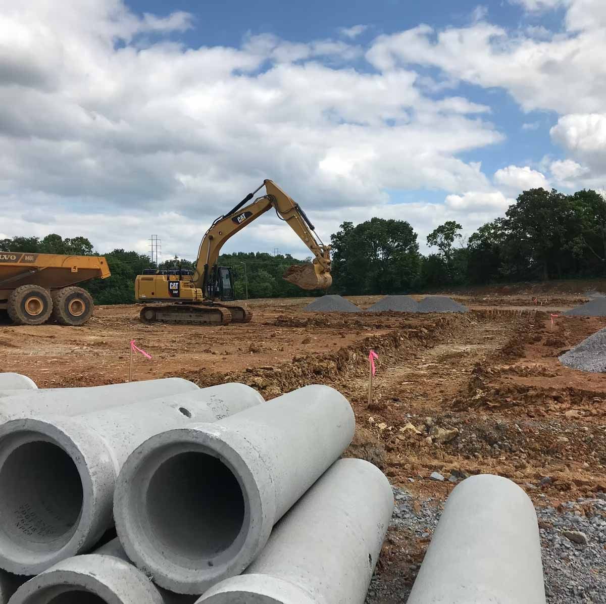Construction site with excavator, dump truck, and concrete pipes; cloudy sky.
