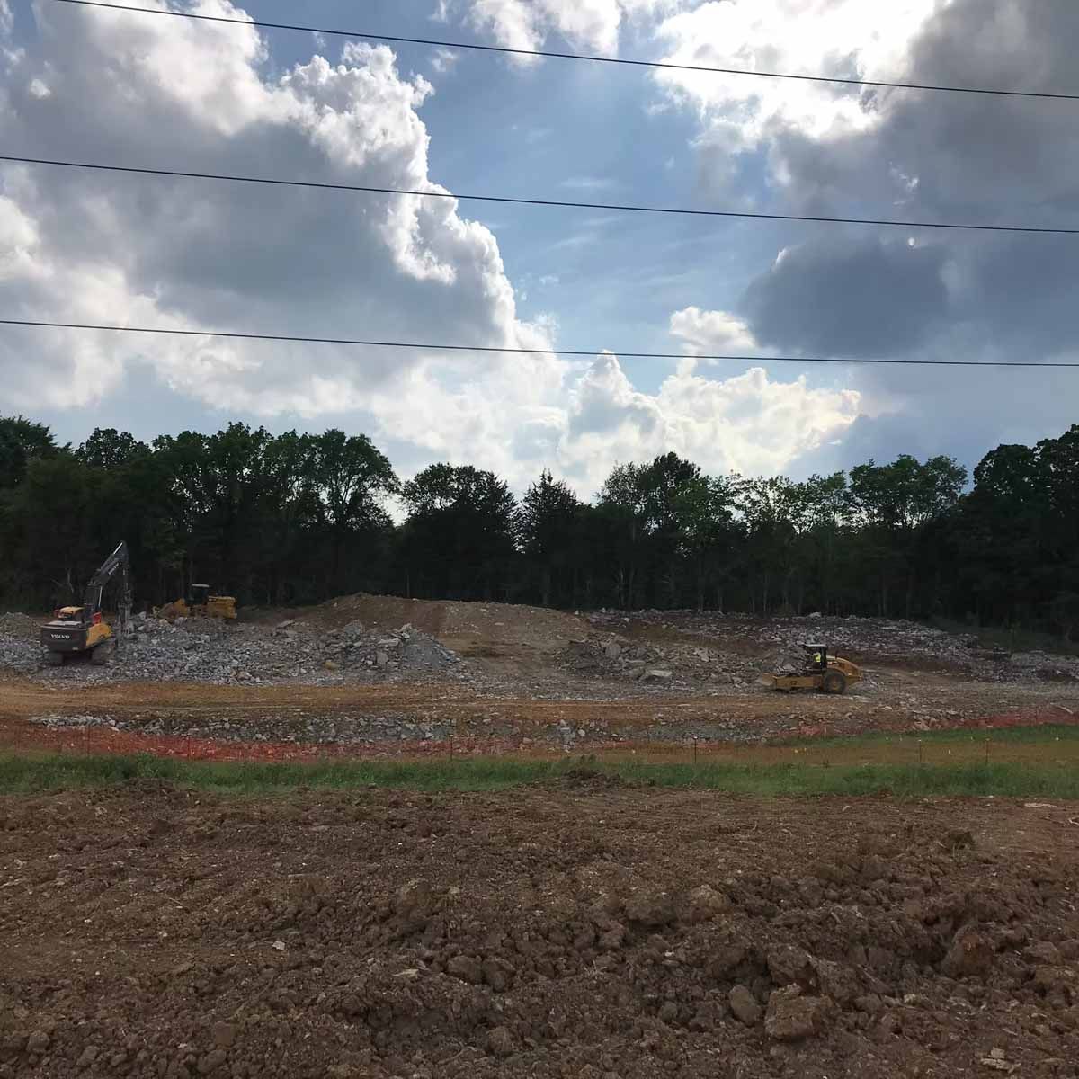 Construction site with heavy machinery, brown earth, rubble pile, and a forest backdrop under a cloudy sky.