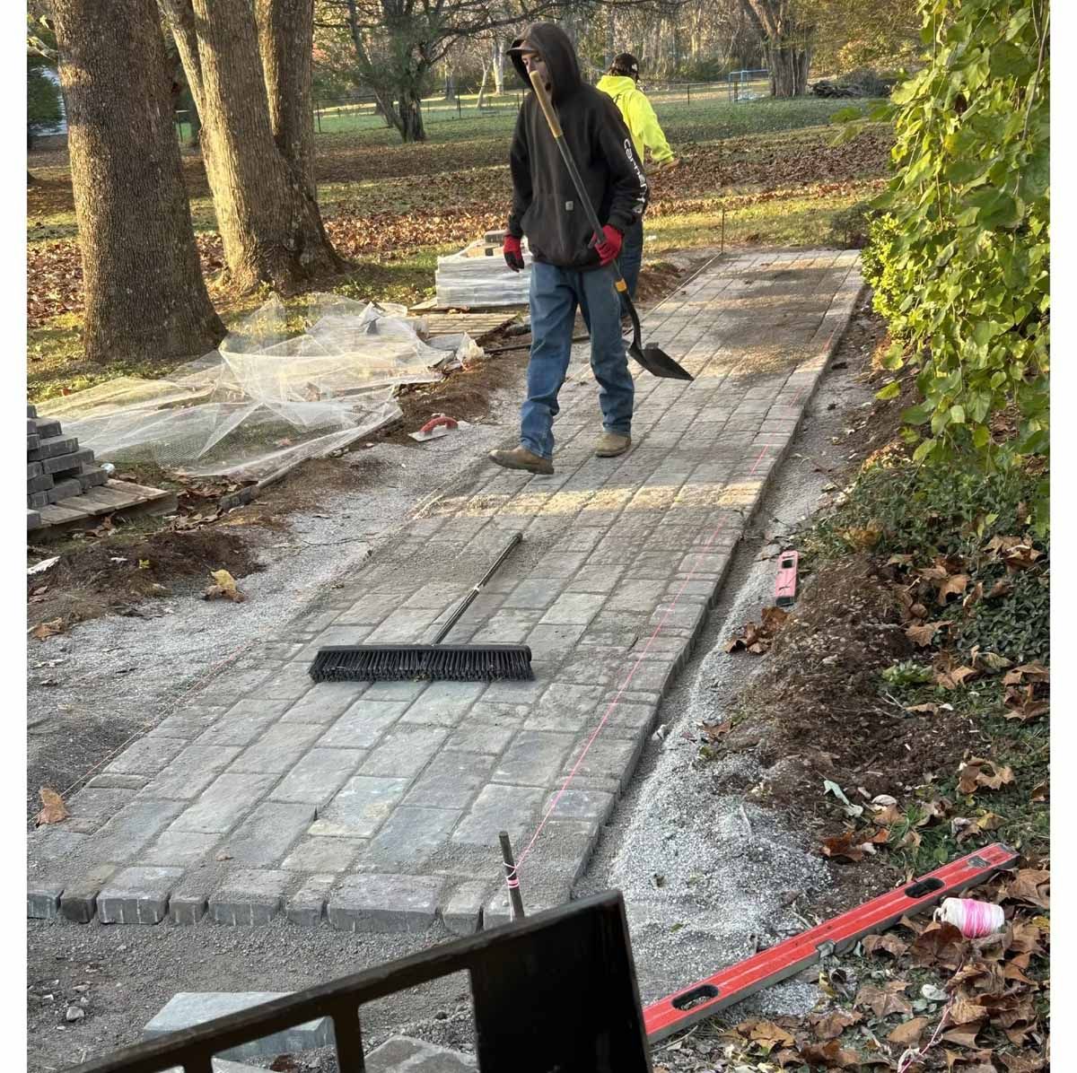 Construction workers laying brick pavers on a pathway. Outdoors, brown trees, gray bricks.