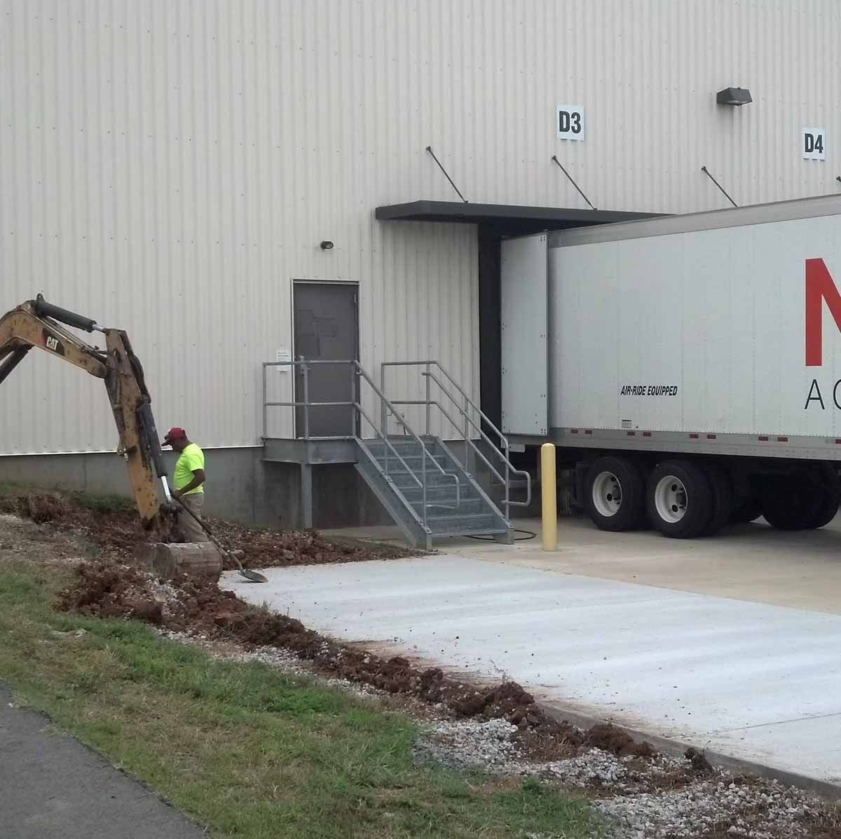 A worker uses an excavator to dig near a loading dock. A truck is parked at the dock.