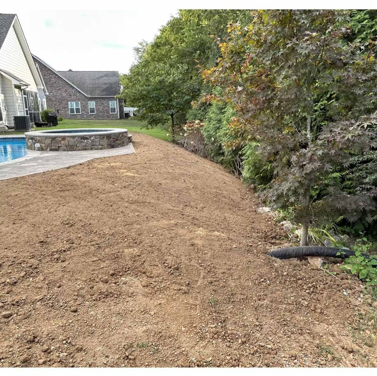 A leveled dirt area next to a retaining wall, beside a pool and house, trees in the background.
