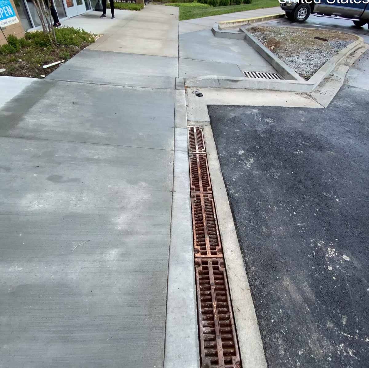 Concrete sidewalk and asphalt road separated by a grated drain. A vehicle is partially visible.