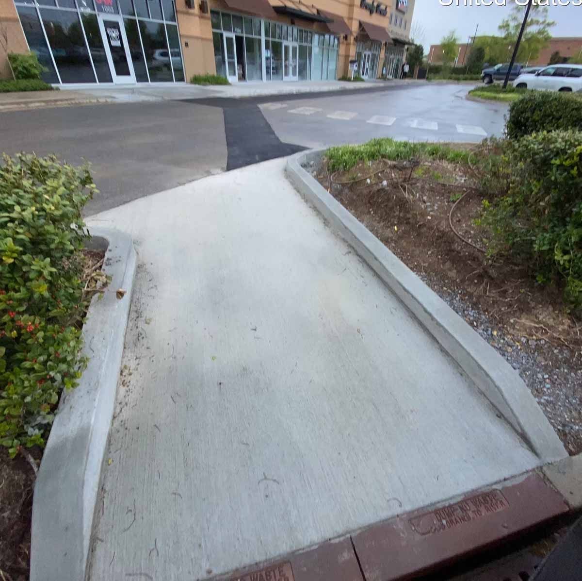 Concrete sidewalk curves towards commercial buildings, lined by bushes and a red brick curb.