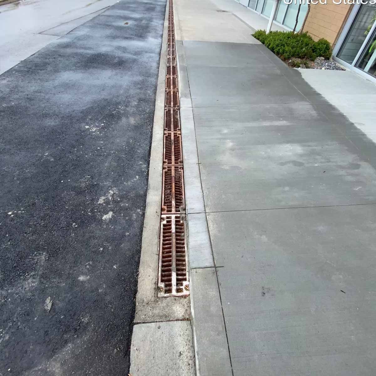 Metal grate drain along a sidewalk and asphalt road next to a building with greenery.