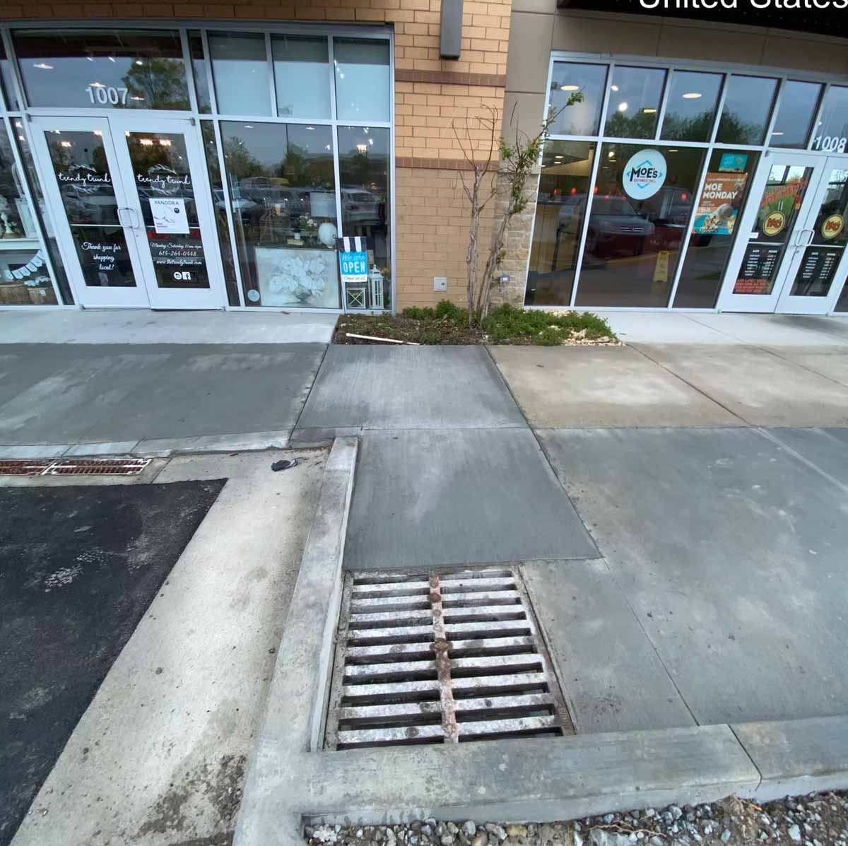 Sidewalk with a grate drain in front of shops with glass windows and doors.