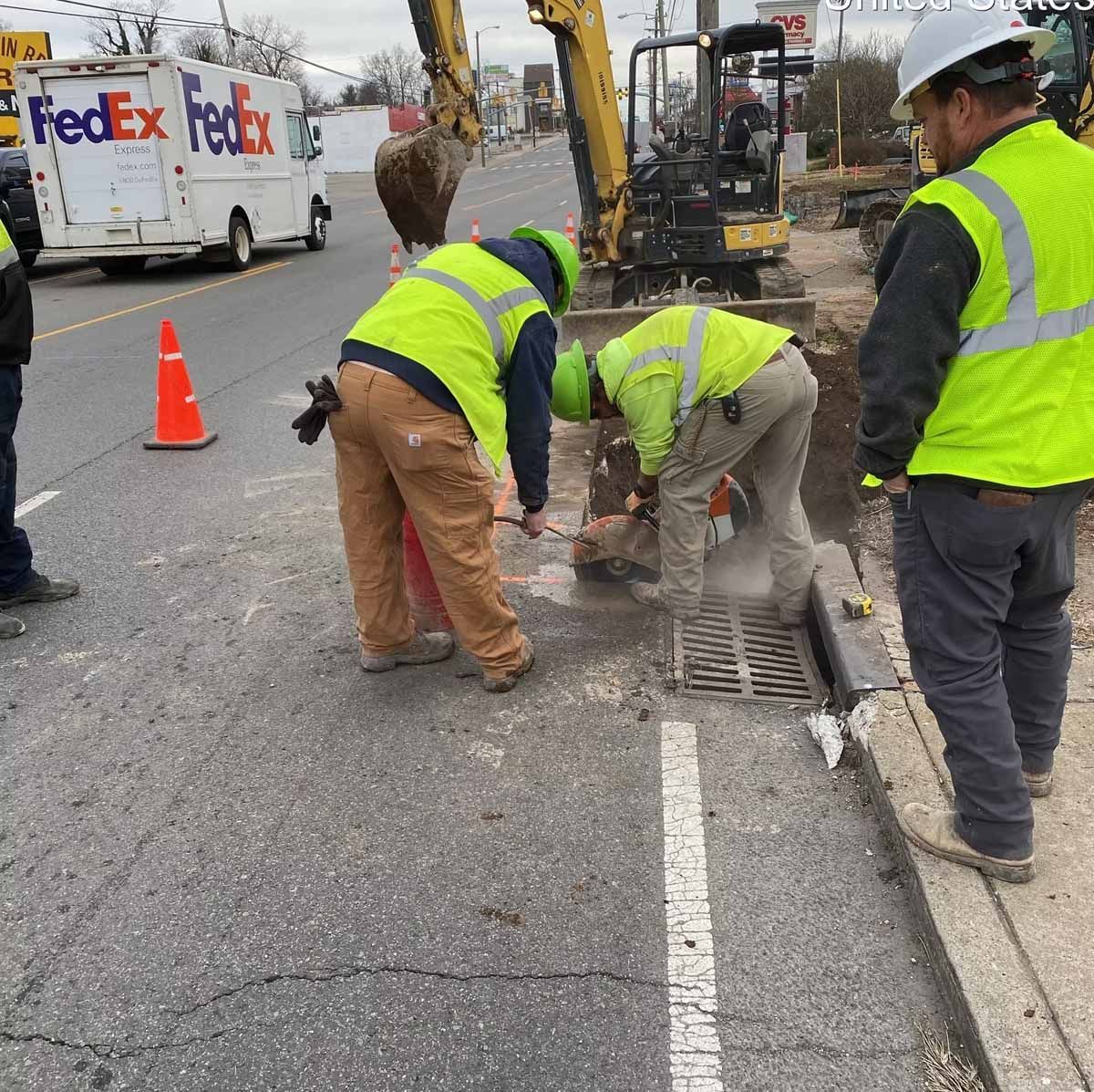 Construction workers cutting into a drain on a street with a FedEx truck and a small excavator.