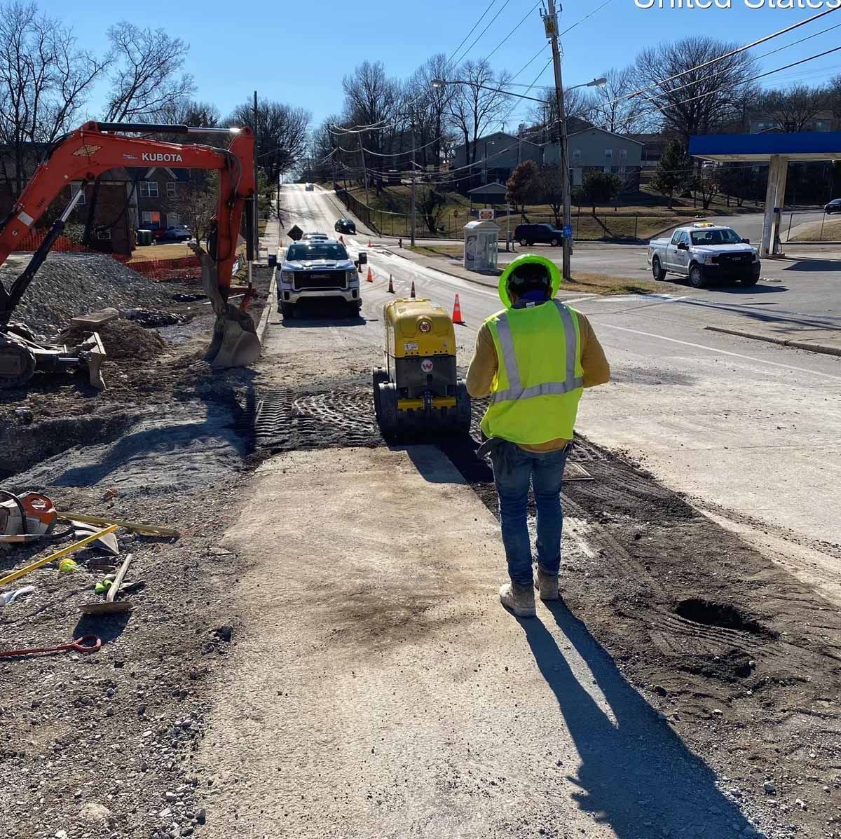 Road construction site. Worker in safety vest operates a compactor machine on a sunny day.