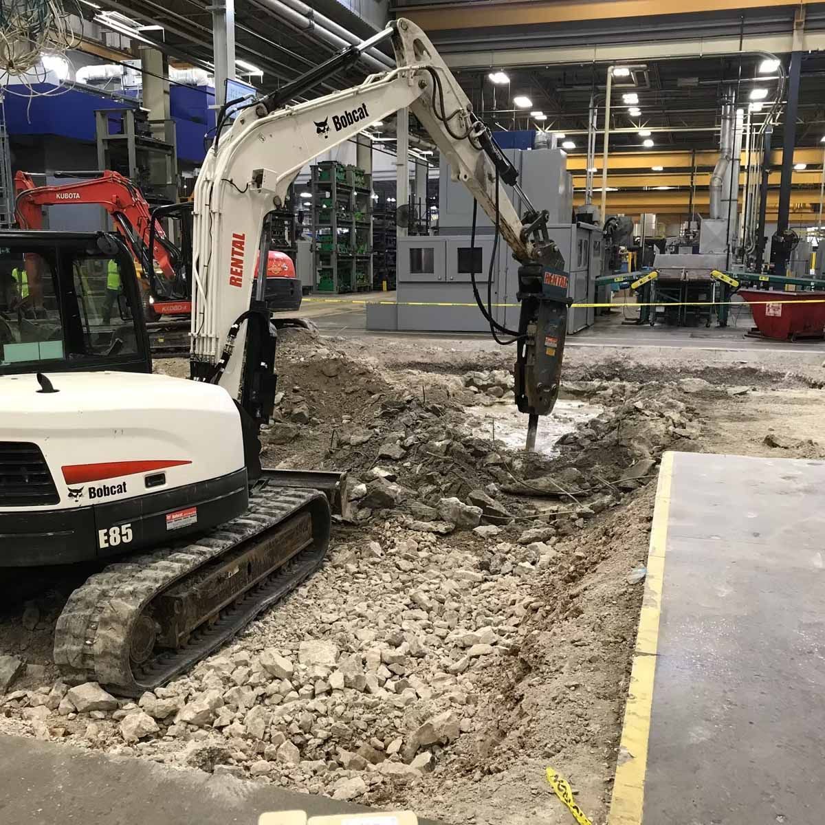 A Bobcat excavator with a jackhammer attachment is breaking up concrete inside a large industrial building.